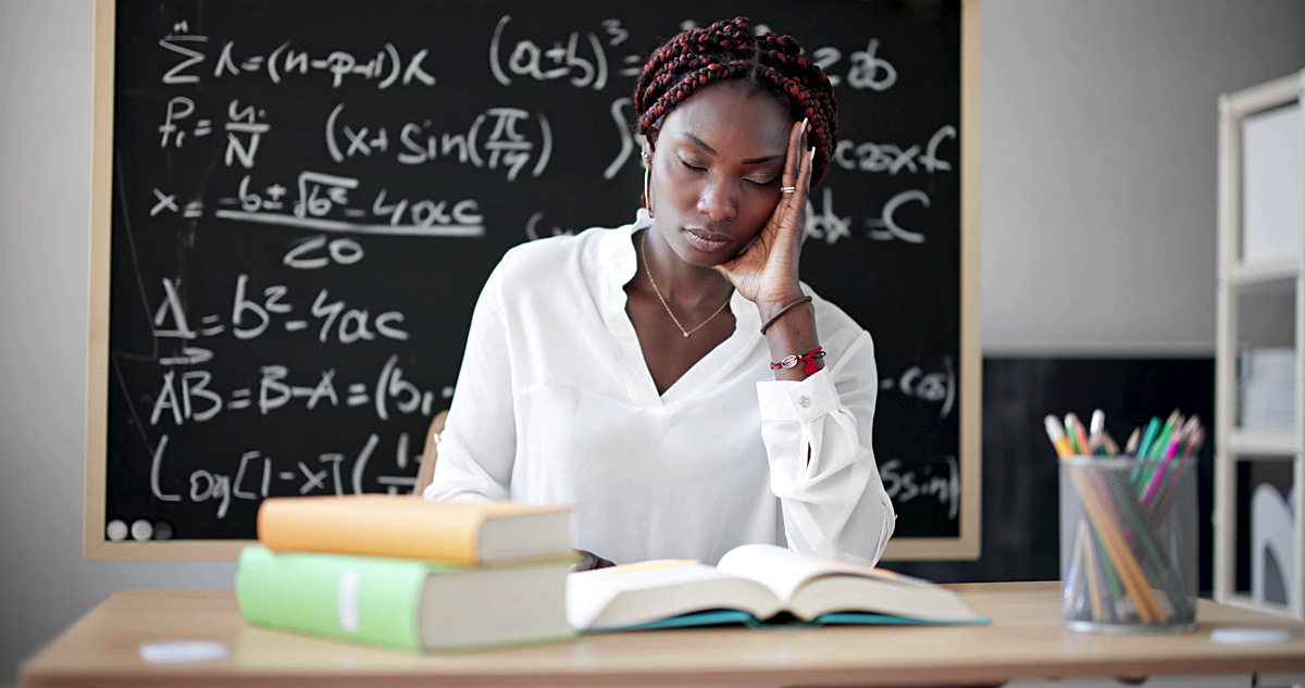 Black woman educator stressed and sitting behind her desk with her eyes closed and head on hand, chalkboard in background with equations
