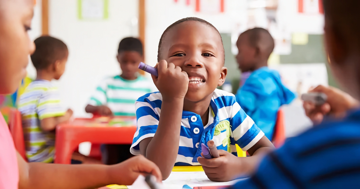 Young Black by sitting at a table with his peers in a classroom coloring with crayons