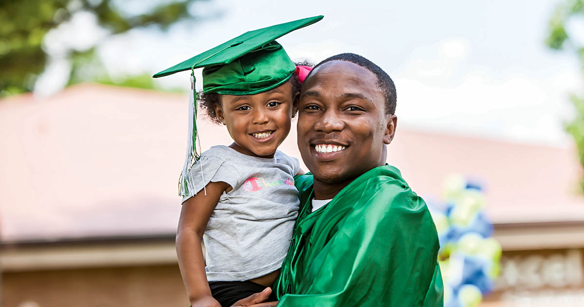 A smiling black adult male graduate holding his child who is wearing his graduation mortar board.