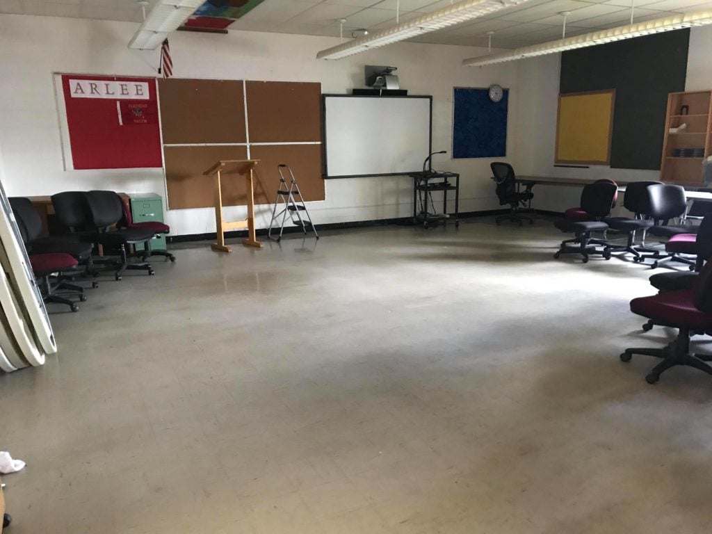An empty classroom with a Smartboard, a bulletin board, a red board that reads"ARLEE, an American Flag, a podium, a step ladder, folded tables, and chairs pushed to the sides of the room.