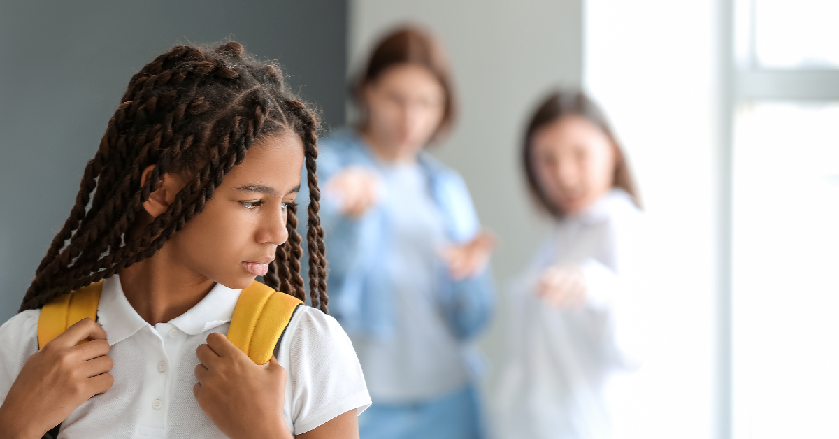 Sad young Black girl with braids wearing yellow backpack in school hallway; Two young white classmates in background