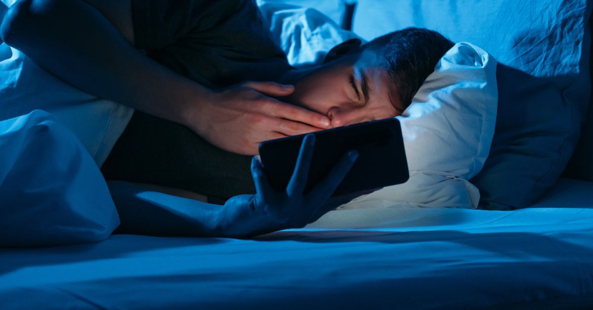 White teenage boy laying in bed in the dark in the glow of his mobile phone, staring at the screen