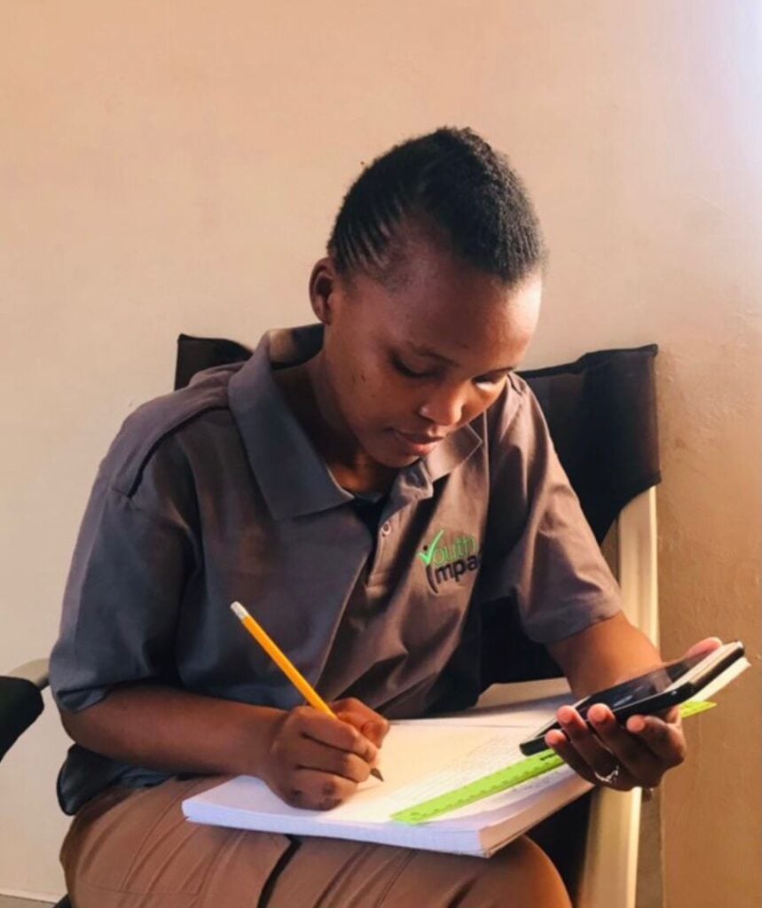 A young Black female student writing with a pencil while holding a calculator and a green ruler. 