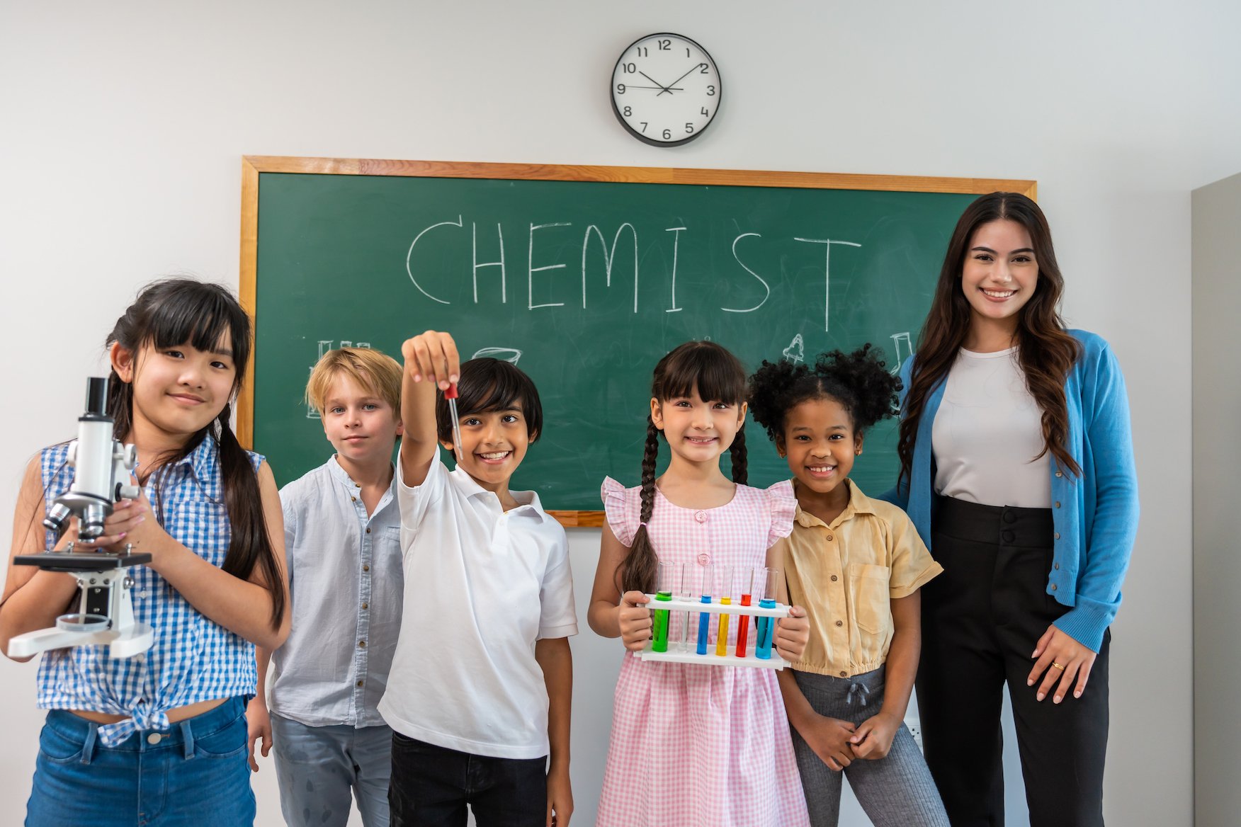 diverse group of school kids with their teacher in front of a chalkboard that says "CHEMIST"